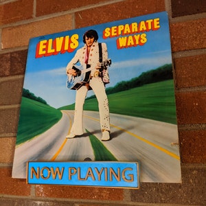 May include: A vintage vinyl record album cover featuring Elvis Presley, with the title "Elvis Separate Ways" in yellow and blue lettering. The album cover shows Elvis standing on a road with a guitar in his hand, wearing a white jumpsuit with a black belt and a black scarf. The album cover is displayed on a brick wall with a blue sign that reads "NOW PLAYING" in gold lettering.