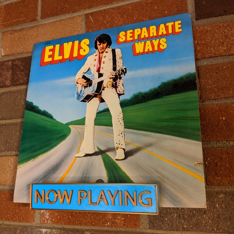 May include: A vintage vinyl record album cover featuring Elvis Presley, with the title "Elvis Separate Ways" in yellow and blue lettering. The album cover shows Elvis standing on a road with a guitar in his hand, wearing a white jumpsuit with a black belt and a black scarf. The album cover is displayed on a brick wall with a blue sign that reads "NOW PLAYING" in gold lettering.