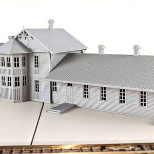 Puede incluir: Una estación de tren modelo gris con un edificio largo y rectangular y una estructura más pequeña, similar a una torre. El edificio tiene un techo a dos aguas y varias ventanas. El modelo está hecho de plástico o madera y está detallado con características realistas.