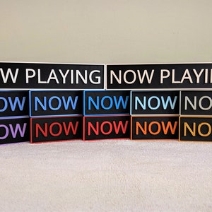 May include: A set of 14 black rectangular signs with white text. The top sign reads "NOW PLAYING". The remaining signs read "NOW" in various colors, including blue, purple, red, orange, and gray.