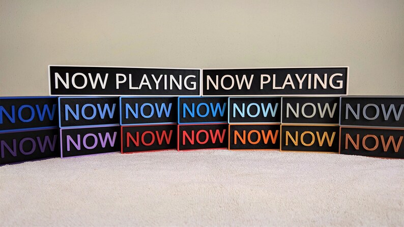 May include: A set of 14 black rectangular signs with white text. The top sign reads "NOW PLAYING". The remaining signs read "NOW" in various colors, including blue, purple, red, orange, and gray.