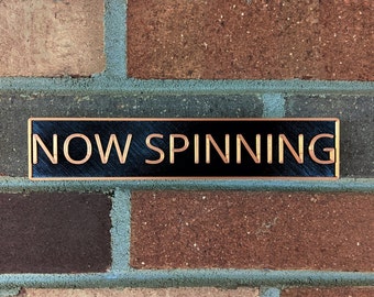 May include: A black sign with a gold border that reads "NOW SPINNING" on a brick wall.
