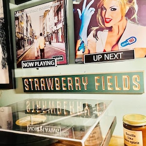 May include: A green sign with gold lettering that reads "STRAWBERRY FIELDS". The sign is mounted on a wall with a record player and two album covers below it. The album covers are for Oasis's "What's the Story Morning Glory?" and Blink-182's "Enema of the State".