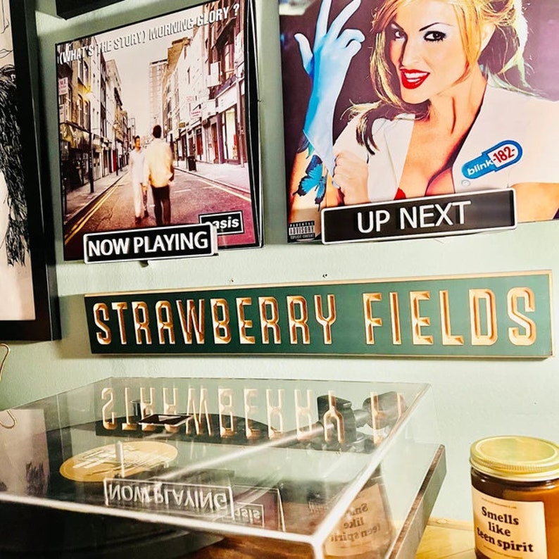 May include: A green sign with gold lettering that reads "STRAWBERRY FIELDS". The sign is mounted on a wall with a record player and two album covers below it. The album covers are for Oasis's "What's the Story Morning Glory?" and Blink-182's "Enema of the State".