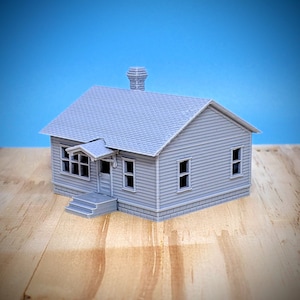 May include: A small, gray model house with a chimney, front porch, and multiple windows. The house has a sloped roof and is made of a ridged material. The model sits on a wooden surface against a blue background.