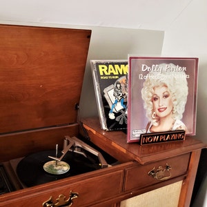 May include: A vintage wooden record player with a turntable and a record on it. The record player has a brown wooden cabinet with a drawer and a door. There are two record covers on top of the cabinet. One record cover is for a Ramones album and the other is for a Dolly Parton album. The Dolly Parton album cover has the text "Dolly Parton 12 of Her Biggest No. 1 Hits" and a sign that says "NOW PLAYING".