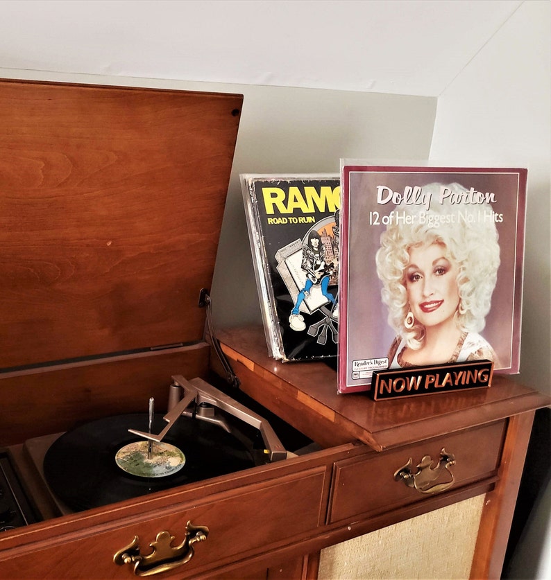 May include: A vintage wooden record player with a turntable and a record on it. The record player has a brown wooden cabinet with a drawer and a door. There are two record covers on top of the cabinet. One record cover is for a Ramones album and the other is for a Dolly Parton album. The Dolly Parton album cover has the text "Dolly Parton 12 of Her Biggest No. 1 Hits" and a sign that says "NOW PLAYING".