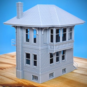 Op de afbeelding: Een grijze schaalmodel van een meerlaags gebouw met een schuin dak, schoorsteen en meerdere ramen. Het gebouw heeft een bakstenen gevel en een klein balkon. Het model staat op een houten oppervlak tegen een blauwe achtergrond.