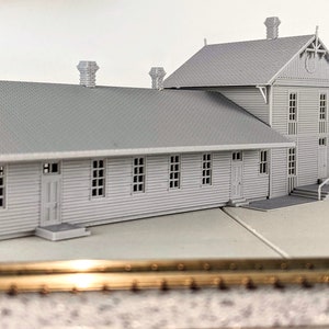 May include: A gray model train station with a long, rectangular building and a smaller, gabled building attached. The station has a platform and a set of tracks in front of it.