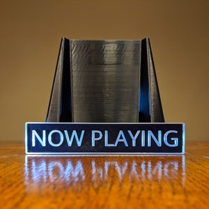 May include: A black plastic stand with a sign that reads "NOW PLAYING" in white letters. The stand is on a wooden surface.