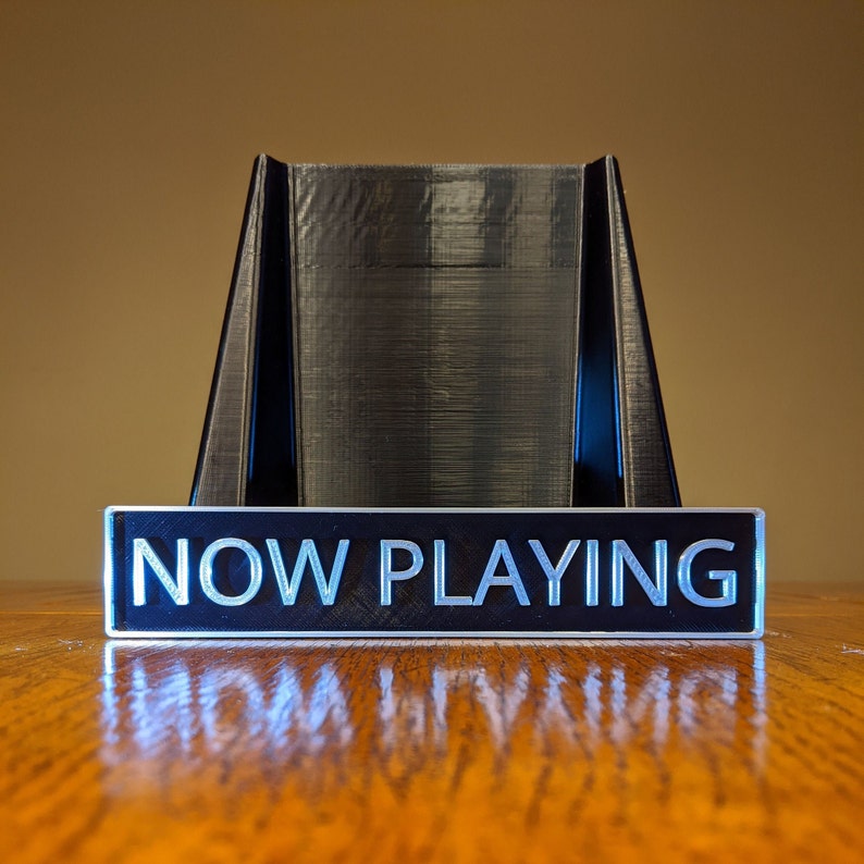 May include: A black plastic stand with a sign that reads "NOW PLAYING" in white letters. The stand is on a wooden surface.