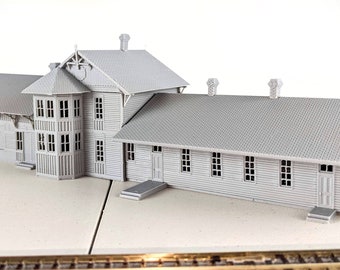 May include: A gray model train station with a long, rectangular building and a smaller, tower-like section. The building has a gabled roof and several windows. The model is made of plastic or wood and is detailed with realistic features.