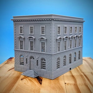 Puede incluir: Maqueta gris de un edificio en miniatura con una fachada detallada, con múltiples ventanas, puertas arqueadas y una textura similar a ladrillos. El modelo se coloca sobre una superficie de madera sobre un fondo azul.