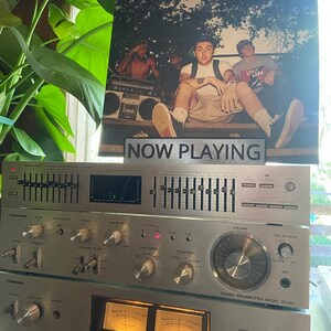 May include: A vintage silver stereo system with a cassette player, equalizer, and amplifier. The system is playing a record with the text "NOW PLAYING" on a sign in front of it. The record cover features three people, one of whom is wearing a shirt with the text "Loose Lips".