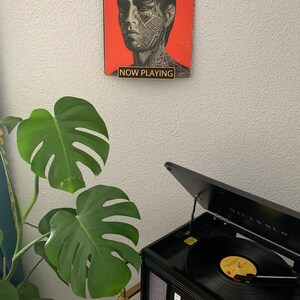 May include: A black record player with a gold record on the turntable. The record player is open and the lid is raised. A green plant is in the background. The image also includes a red poster with a black and white image of a person with tattoos. The poster has the text "NOW PLAYING" on it.