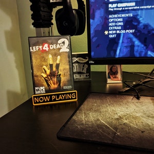 May include: A black and yellow "Now Playing" sign with the video game case for Left 4 Dead 2 standing in front of it. The game case features a zombie hand reaching out from the cover art.
