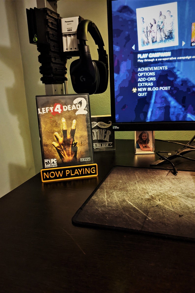 May include: A black and yellow "Now Playing" sign with the video game case for Left 4 Dead 2 standing in front of it. The game case features a zombie hand reaching out from the cover art.