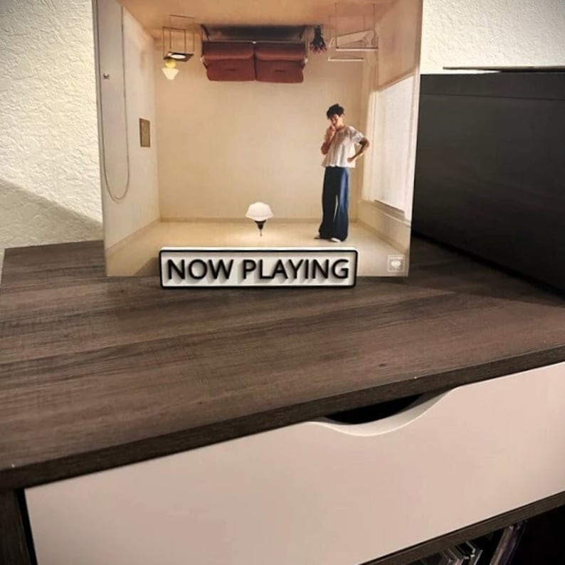 May include: A white sign with black text that reads "NOW PLAYING" on a brown wooden surface.