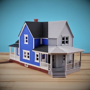 May include: A split-view model house featuring a blue exterior with white trim on the left side and a gray, unpainted exterior on the right side. The structure includes a black roof, a front porch with railings, and multiple windows.