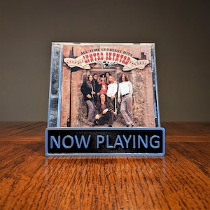 May include: A silver metal sign with the text "NOW PLAYING" in black letters. The sign is in front of a CD case with a photo of a band called "Lynyrd Skynyrd" on the cover. The CD case has the text "All Time Greatest Hits" on the front.