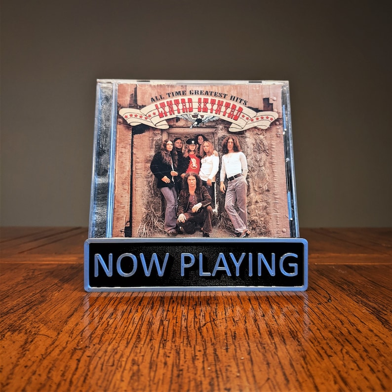 May include: A silver metal sign with the text "NOW PLAYING" in black letters. The sign is in front of a CD case with a photo of a band called "Lynyrd Skynyrd" on the cover. The CD case has the text "All Time Greatest Hits" on the front.
