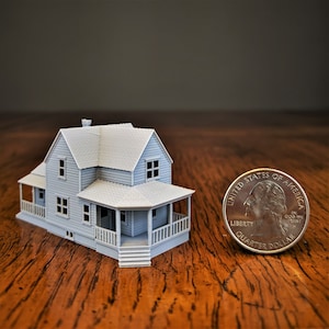 May include: A miniature gray house model featuring a gabled roof, siding texture, and a wrap-around porch with railings. The scale of the structure is shown next to a United States quarter dollar coin for size comparison on a wooden surface.