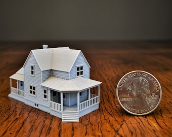 May include: A miniature gray house model featuring a gabled roof, siding texture, and a wrap-around porch with railings. The scale of the structure is shown next to a United States quarter dollar coin for size comparison on a wooden surface.