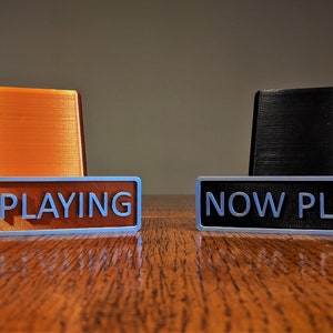May include: Two "NOW PLAYING" signs, one with an orange base and one with a black base. The signs are made of a silver metal with white lettering.
