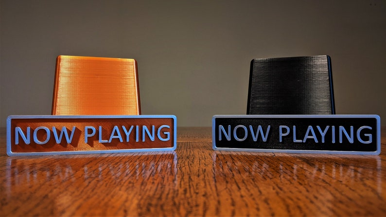 May include: Two "NOW PLAYING" signs, one with an orange base and one with a black base. The signs are made of a silver metal with white lettering.