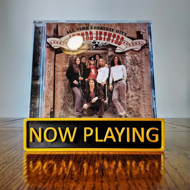 May include: A yellow sign with the text "NOW PLAYING" in black letters, in front of a vintage record album cover featuring the band "Lynyrd Skynyrd" with the text "All Time Greatest Hits".
