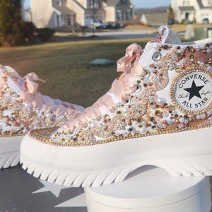 May include: White high-top Converse sneakers with a platform sole, embellished with gold and pink rhinestones, pearls, and a pink satin ribbon. The Converse logo is visible on the side of the shoe.