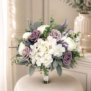 May include: A bridal bouquet featuring white hydrangeas, lavender roses, and sprigs of greenery. The arrangement is tied with a white ribbon and sits on a round, beige ottoman. The bouquet includes white peonies and lavender accents.
