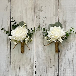 May include: Two white rose boutonnieres with greenery and white baby's breath, tied with twine.