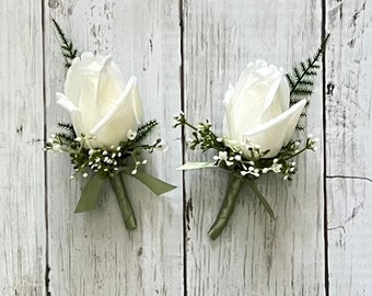 May include: Two white rose boutonnieres with green foliage and a green ribbon.