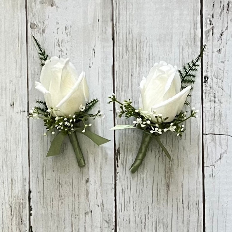 May include: Two white rose buttonholes with green foliage and a green ribbon.