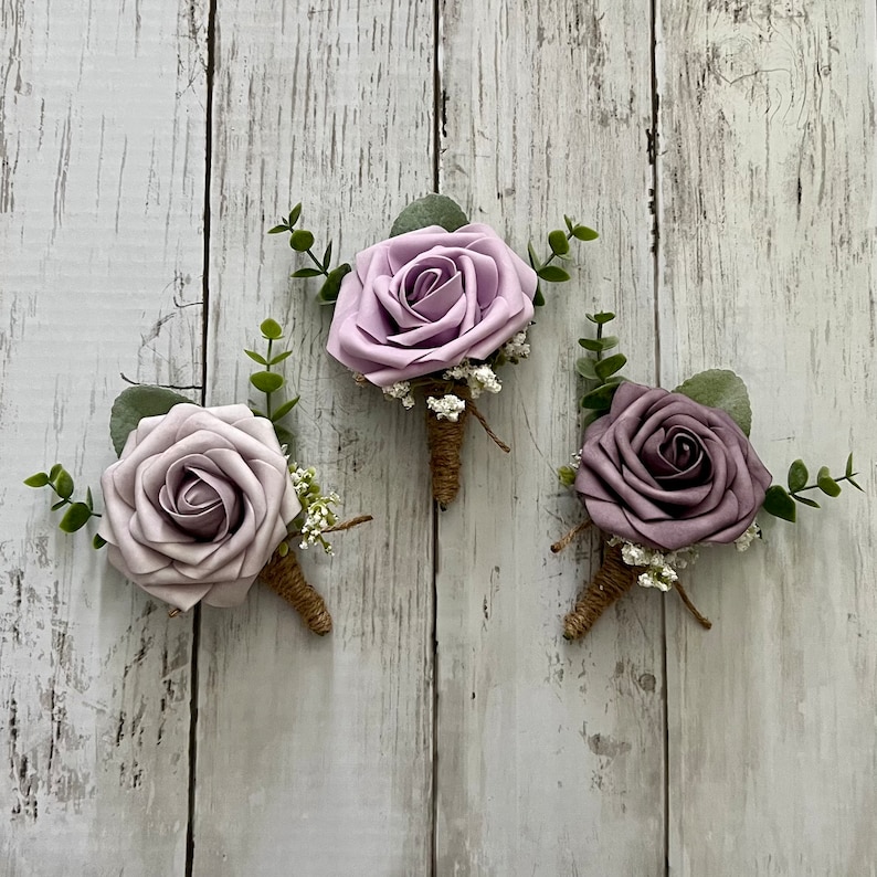 Op de afbeelding: Drie florale corsages met enkele rozen in tinten lichtpaars, lavendel en stoffig mauve. Elke roos is geaccentueerd met kleine groene eucalyptusblaadjes, piepkleine witte vulbloemen en een steel die is omwikkeld met bruin touw.