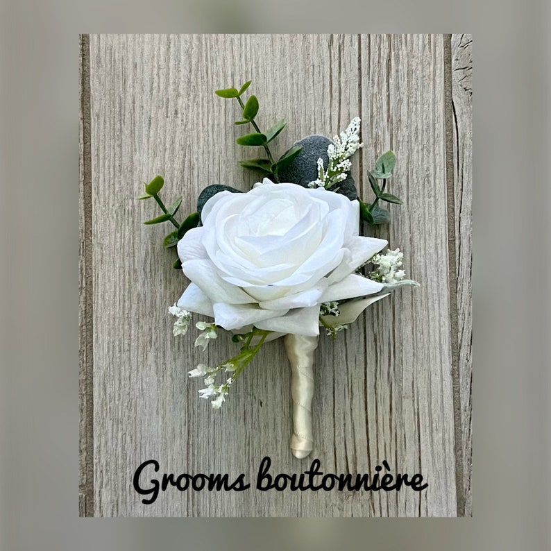 May include: A white rose boutonniere with greenery and white baby's breath. The boutonniere is tied with a white satin ribbon and has the text "Grooms boutonni&egrave;re" written below it.