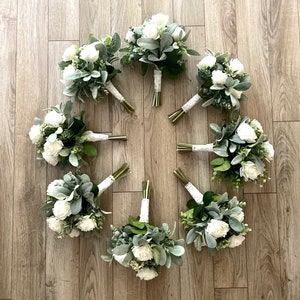 K&ouml;nnte beinhalten: Acht Blumenstr&auml;u&szlig;e in kreisf&ouml;rmiger Anordnung, bestehend aus wei&szlig;en Rosen, gr&uuml;nen Eukalyptusbl&auml;ttern und weichem, grauem Woll-Ziest. Die Stiele jedes Strau&szlig;es sind mit einem wei&szlig;en Spitzenband zusammengebunden, was ein einheitliches Set ergibt.