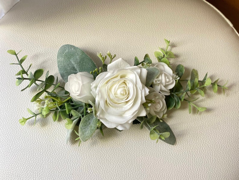 K&ouml;nnte beinhalten: Ein Blumenarrangement mit mehreren wei&szlig;en Rosen in verschiedenen Gr&ouml;&szlig;en, akzentuiert durch gr&uuml;ne Eukalyptusbl&auml;tter und kleine Zweige. Die Komposition ist horizontal ausgerichtet und zeigt geschichtete Bl&uuml;tenbl&auml;tter sowie weiches, mattgr&uuml;nes Blattwerk.