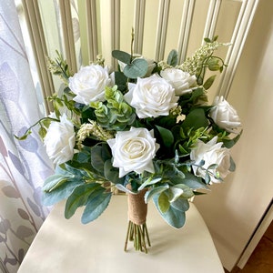 K&ouml;nnte beinhalten: Ein Blumenstrau&szlig; mit wei&szlig;en Rosen und verschiedenen gr&uuml;nen Blattpflanzen, darunter Eukalyptus und Woll-Ziest-Bl&auml;tter. Die Stiele sind zusammengebunden und mit einem strukturierten braunen Juteband umwickelt, was ein geordnetes Blumenarrangement ergibt.