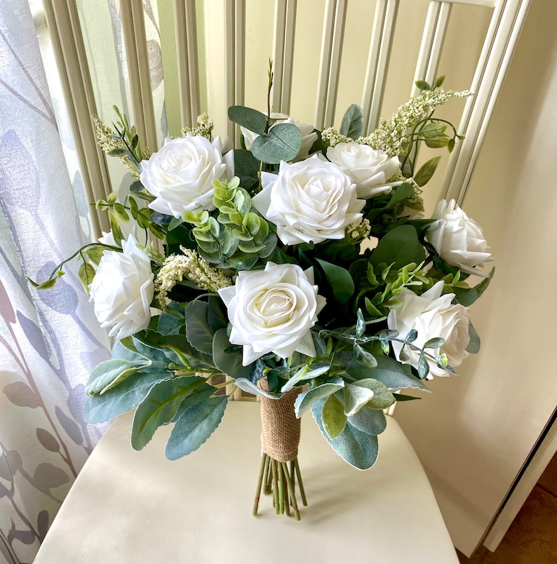 K&ouml;nnte beinhalten: Ein Blumenstrau&szlig; mit wei&szlig;en Rosen und verschiedenen gr&uuml;nen Blattpflanzen, darunter Eukalyptus und Woll-Ziest-Bl&auml;tter. Die Stiele sind zusammengebunden und mit einem strukturierten braunen Juteband umwickelt, was ein geordnetes Blumenarrangement ergibt.