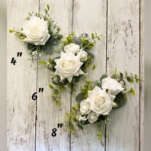K&ouml;nnte beinhalten: Drei Blumenarrangements mit wei&szlig;en Rosen, gr&uuml;nen Eukalyptusbl&auml;ttern und kleinen wei&szlig;en F&uuml;llblumen. Die Arrangements sind mit Gr&ouml;&szlig;en von 10 cm, 15 cm und 20 cm beschriftet, was die L&auml;nge des jeweiligen floralen Einzelst&uuml;cks angibt.