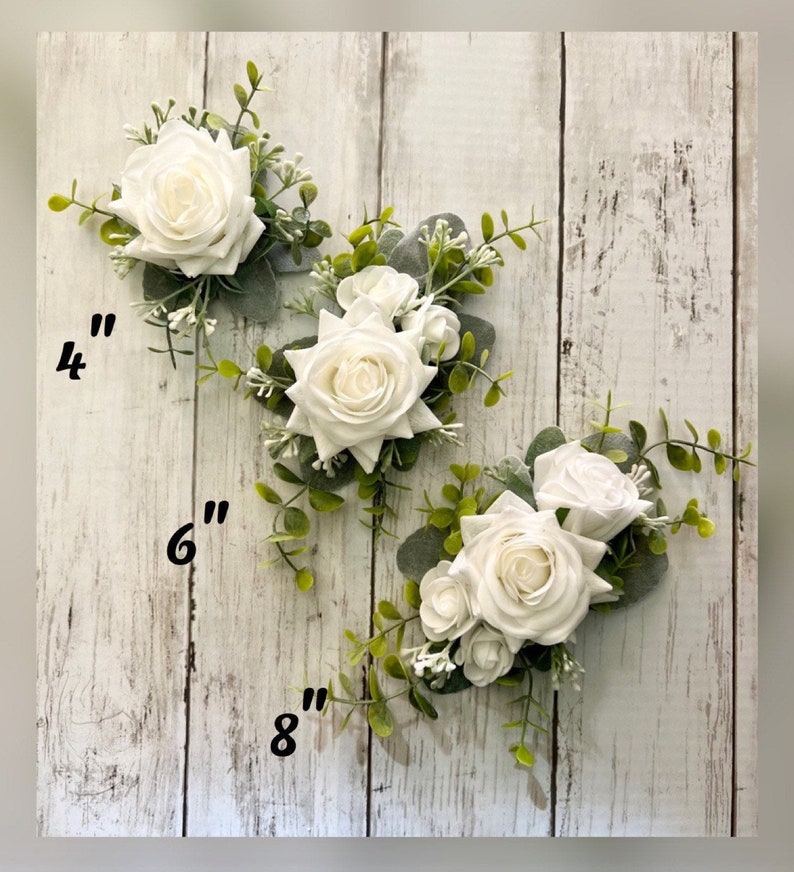 K&ouml;nnte beinhalten: Drei Blumenarrangements mit wei&szlig;en Rosen, gr&uuml;nen Eukalyptusbl&auml;ttern und kleinen wei&szlig;en F&uuml;llblumen. Die Arrangements sind mit Gr&ouml;&szlig;en von 10 cm, 15 cm und 20 cm beschriftet, was die L&auml;nge des jeweiligen floralen Einzelst&uuml;cks angibt.