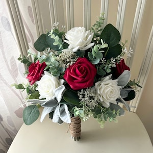 May include: A bridal bouquet featuring red and white roses, accented with green eucalyptus and silver foliage. The stems are wrapped in twine. The arrangement is set against a neutral background, suggesting a wedding or event theme.