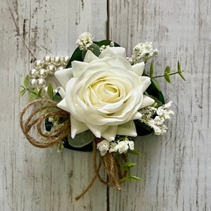 May include: A floral corsage featuring a large white rose as the central element, accented with small white filler flowers, green leaves, a strand of white faux pearls, and loops of natural brown twine arranged in a decorative cluster.