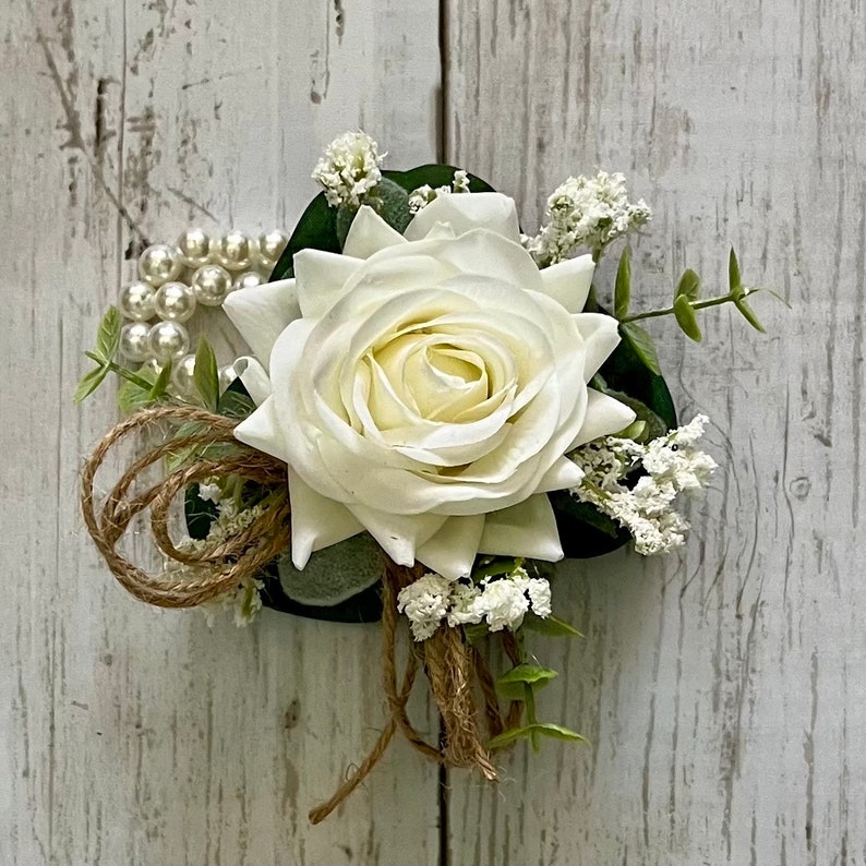May include: A floral corsage featuring a large white rose as the central element, accented with small white filler flowers, green leaves, a strand of white faux pearls, and loops of natural brown twine arranged in a decorative cluster.