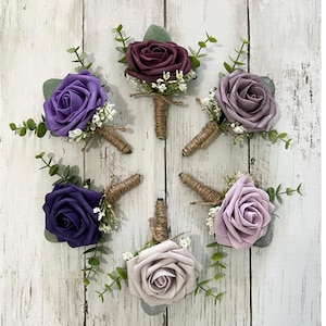 May include: Six small bouquets of artificial flowers, each with a single rose and greenery, tied with twine. The roses are in shades of purple, lavender, and burgundy.