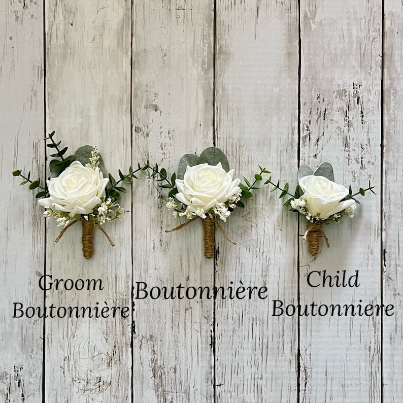 May include: Three boutonni&egrave;res featuring white roses, green eucalyptus leaves, and small white filler flowers. Each stem is wrapped in brown twine. The text Groom Boutonni&egrave;re, Boutonni&egrave;re, and Child Boutonni&egrave;re is printed below each respective item.
