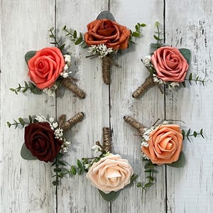 Op de afbeelding: Zes kleine bloemstukken met perzik-, koraal- en bordeauxrode rozen, witte baby's adem en eucalyptus takjes. Elk arrangement is vastgebonden met touw.