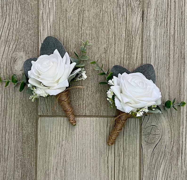 May include: Two white rose boutonnieres with greenery and twine accents. The roses are made of fabric and the greenery is made of plastic.